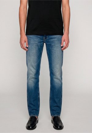 Uomo con jeans blu slavati, scarpe nere slip-on e una t-shirt nera, in piedi davanti a uno sfondo chiaro e uniforme.
