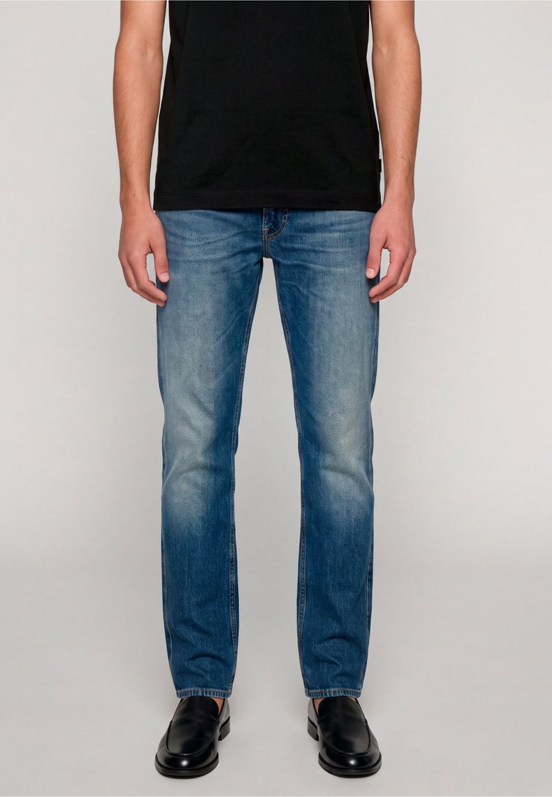 Homme portant un jean bleu délavé, des chaussures noires sans lacets et un t-shirt noir, debout devant un fond clair uni.
