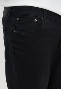 Μαύρο τζιν με κοντινή όψη στην μπροστινή τσέπη, που τονίζει την υφή, τις ραφές και τις λεπτομέρειες με μεταλλικό κουμπί.