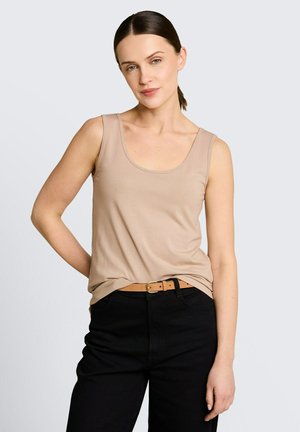 Femme aux cheveux bruns attachés en queue de cheval, portant un haut beige sans manches rentré dans un pantalon noir taille haute avec une ceinture couleur camel, debout devant un fond blanc.