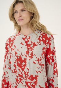 Blusa con motivo rosso e beige, scollatura tonda, chiusura con bottoni, maniche lunghe e una texture morbida. Presenta una tasca sul davanti.