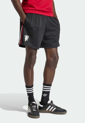 Mand iført sorte atletikshorts med røde og hvide striber, sorte Adidas-strømper med hvide striber og sorte Adidas-sneakers med hvide striber.