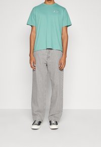 Osoba v mátově zeleném tričku Levi's, světle šedých volných džínech a černých nízkých teniskách stojící na bílém pozadí.