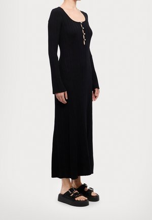 Woman wearing a long black ribbed dress with front buttons and black platform sandals with double buckles, standing against a plain background.