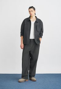 Tamsiai pilkas marškiniai su kišenėmis, dėvimi virš baltos marškinėlių, derinami su laisvais pilkais kelnais ir šviesiai pilkomis avalynėmis ant mėlyno fono.