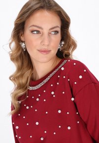 Bordó pulóver, mely szegecses díszítéssel rendelkezik, különböző méretű gyöngyökkel és gyöngyfűzött nyakkivágással, sima textúrával és kényelmes, laza fazonnal.