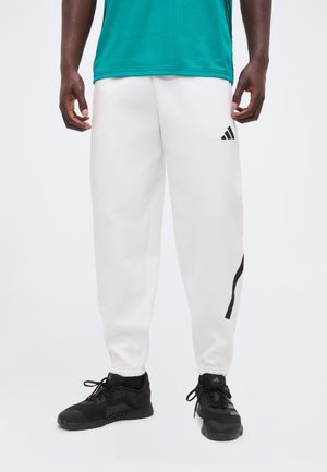 Mężczyzna ubrany w białe spodnie dresowe Adidas z czarnym logo i czarne sneakersy, stojący na jednolitym tle.