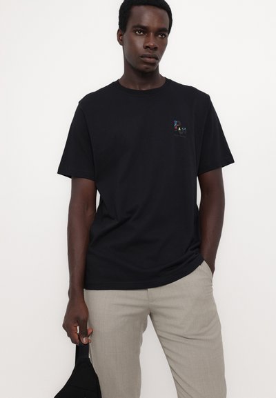 Tricou din bumbac negru cu mâneci scurte, având un logo mic și colorat pe piept, asortat cu pantaloni gri deschis și o geantă neagră.