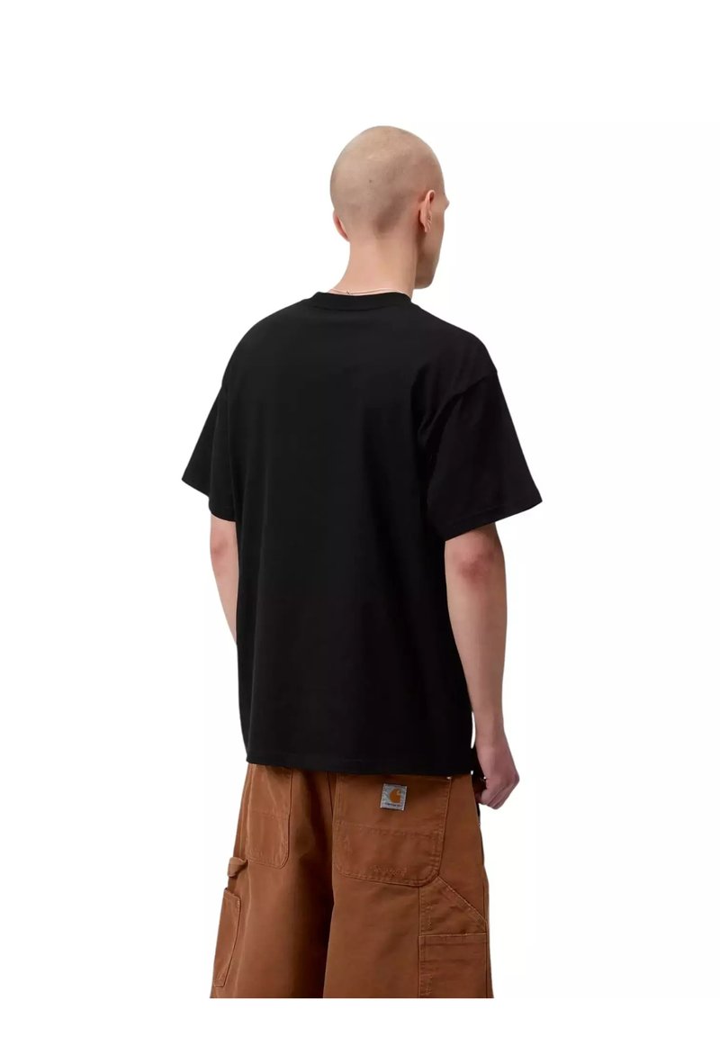 Mann mit rasiertem Kopf, der ein schwarzes T-Shirt und braune Cargo-Shorts trägt, steht mit dem Rücken zu einem weißen Hintergrund.