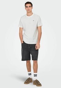 White cotton t-shirt with a small black logo, paired with dark denim shorts. Model wears striped socks and brown lace-up shoes.