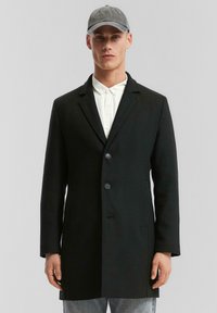 Schwarzer Wollmantel mit taillierter Passform, einreihigem Design, zwei vorderen Knöpfen und seitlichen Taschen. Über einem weißen Hemd getragen und mit einer grauen Mütze kombiniert.
