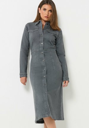 Femme portant une robe en jean grise cintrée à manches longues avec des boutons, debout devant un fond blanc uni.