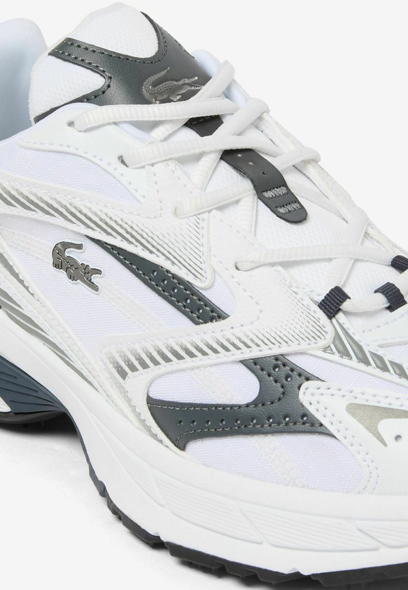 Weißer und grauer Sportschuh mit Schnürverschluss, strukturierter Sohle und einem kleinen Krokodil-Logo an der Seite und auf der Zunge.