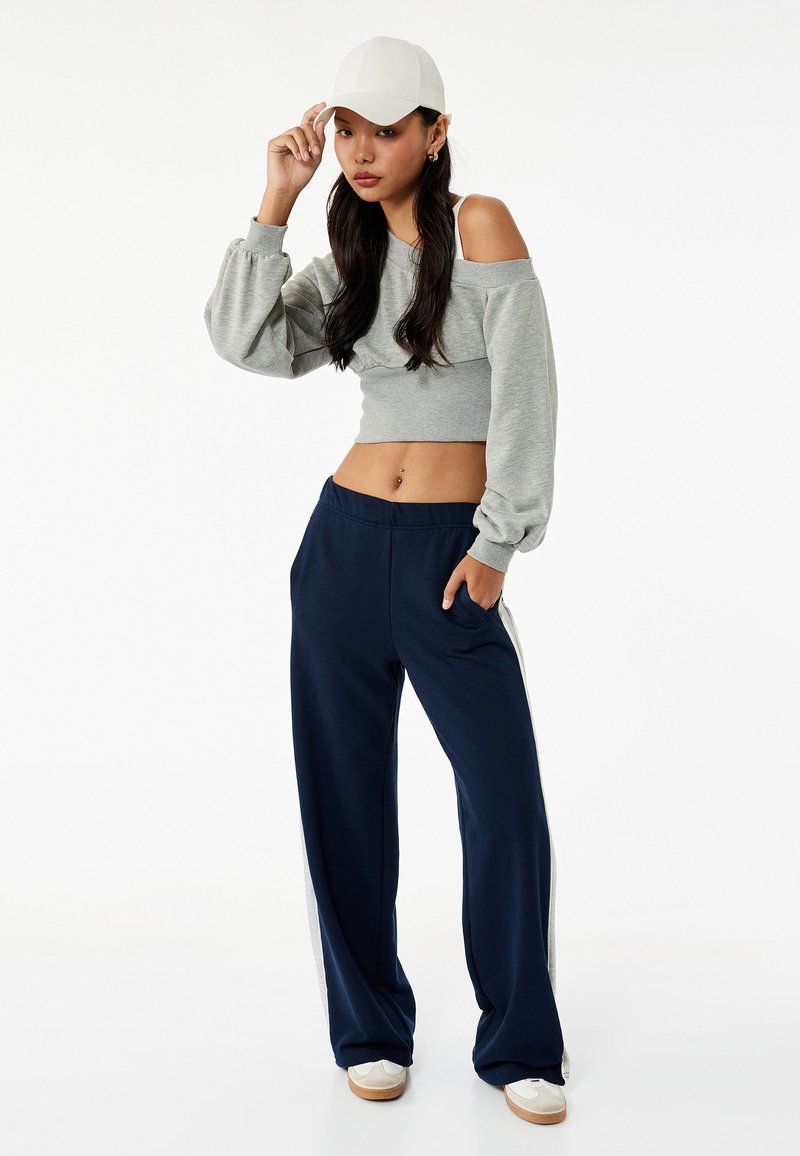 Grijze cropped sweatshirt met ballonmouwen, gecombineerd met marineblauwe wijde broek met een witte streep. Witte pet en schoenen maken de look compleet.