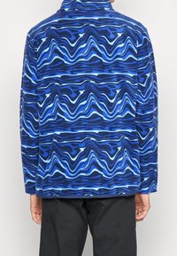 Giacca in pile caratterizzata da un motivo ondulato blu con tonalità diverse di blu e bianco, progettata con colletto alto e maniche lunghe.