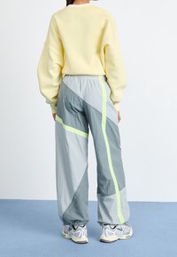 Person i pastellgul sweatshirt, tvåtoniga gråbyxor med neonstripes och vita sneakers står på blått golv, vänd bort.