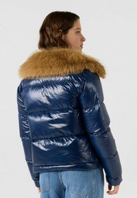 Donzen jack in marineblauw met glanzende textuur, voorzien van een kraag van imitatiebont in lichtbruin. Het ontwerp bevat gewatteerde delen en een aansluitend silhouet.