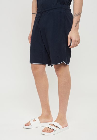 Pantaloni corti blu navy con un motivo testurizzato, cintura elastica e bordo bianco. Abbinati a sandali bianchi con due cinturini regolabili.