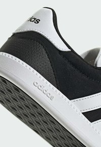 Zapato deportivo en blanco y negro con parte superior sintética texturizada, detalles en cuero liso y suela de goma con patrón geométrico de la banda de rodadura.