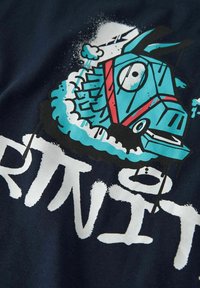 Design grafico blu di una testa stilizzata di animale su tessuto scuro; presenta contorni marcati, schizzi di vernice e testo bianco sotto.