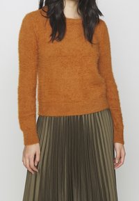 Donna che indossa un maglione fuzzy arancione ruggine con maniche lunghe e una gonna plissettata verde oliva, in piedi di fronte a uno sfondo chiaro e semplice.