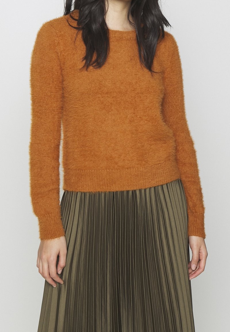 Donna che indossa un maglione fuzzy arancione ruggine con maniche lunghe e una gonna plissettata verde oliva, in piedi di fronte a uno sfondo chiaro e semplice.
