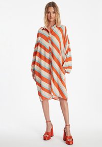 Robe oversize à col, en orange, crème et rayures diagonales bleues ; dotée d'une fermeture à boutons et de manches amples ; portée avec des talons plateforme.