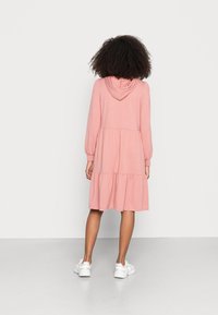 Roze hoodie-jurk met lange mouwen, gelaagde rok, geribbelde manchetten en een zachte textuur, gecombineerd met witte sneakers op een effen achtergrond.