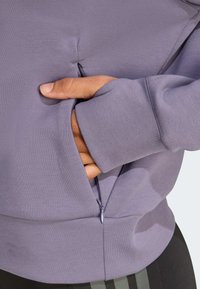 Felpa viola con tasca laterale con zip e polsini a coste, realizzata in un tessuto morbido e liscio con un design aderente.