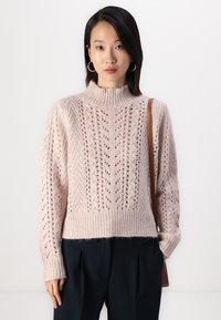 Világosszürke kötött pulóver magas nyakkal, nyitott mintával és bordázott aljjal. Sötét nadrággal és barna válltáskával viselve.