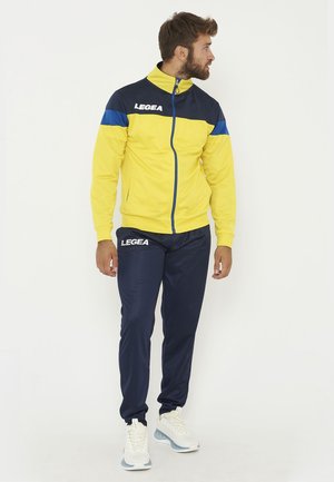 Homme portant une veste de sport jaune et bleu marine et un pantalon de sport bleu marine, regardant vers sa droite, debout devant un fond blanc.