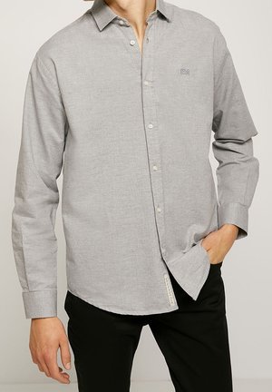 Homme portant une chemise à manches longues gris clair boutonnée avec un pantalon noir, une main dans la poche et l’autre détendue à ses côtés.