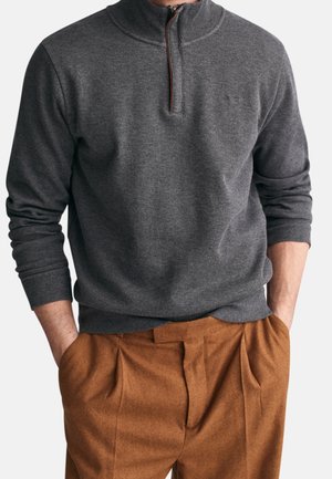 Mann trägt dunkelgrauen Viertel-Zip-Pullover mit gerippten Bündchen und Kragen, kombiniert mit hochgeschnittenen braunen Faltenhosen, Hände in den Taschen.
