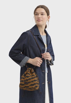 Mujer con un abrigo de mezclilla y una camisa a rayas que lleva un bolso de punto marrón y negro con cordón colgado del hombro.