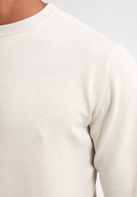 Pulover crem deschis cu guler rotund ribcat, având un model texturat de tricot și un material neted. Detaliile includ cusături subtile pe umăr.