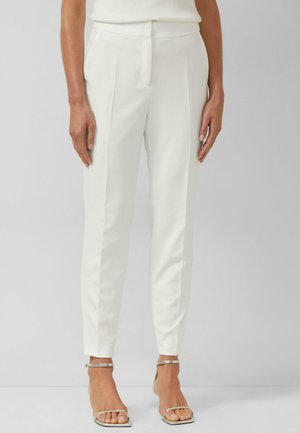 Frau trägt weiße maßgeschneiderte Hose und silberne offene Sandaletten mit Knöchelriemen, steht vor einem schlichten hellen Hintergrund.