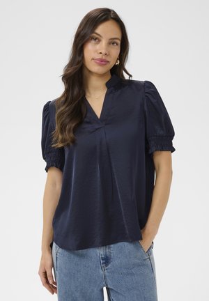 Femme aux longs cheveux foncés portant un chemisier bleu marine à manches courtes bouffantes et un jean bleu clair, debout avec une main dans la poche.