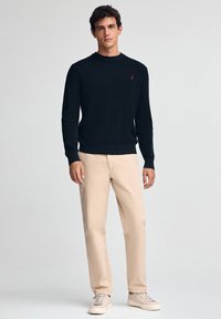 Mann trägt einen schwarzen Langarmpullover, beige Hosen und beige Sneaker und steht vor einem schlichten hellen Hintergrund.