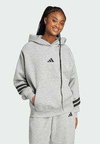 Siva kapuljača s kenguruh džepom. Ima crne trostruke akcentne trake na rukavima i mali crni Adidas logo. Mekana tekstura.