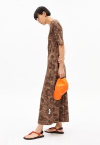 Brown tie-dye maxi dress with short sleeves, side slits, and a discreet tag. Complemented by a bright orange cap and black sandals.