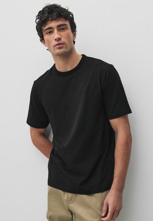 Mann mit lockigem dunklem Haar, der ein schlichtes schwarzes T-Shirt mit kurzen Ärmeln und beige Hosen trägt und vor einem weißen Hintergrund steht.