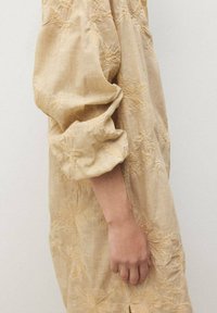 Abito beige ricamato con maniche lunghe e voluminose, caratterizzato da motivi floreali, polsini arricciati e una vestibilità morbida. Tessuto liscio e testurizzato.