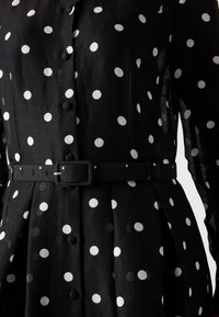Abito nero con pois bianchi, maniche lunghe, abbottonatura frontale, vita con cintura e gonna plissettata. Realizzato in un tessuto leggero e testurizzato.