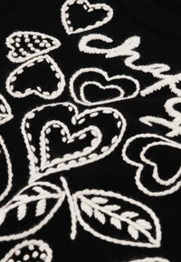 Zwarte trui met witte geborduurde harten en bladeren, met verschillende patronen en texturen. Opvallende stikdetails accentueren het ontwerp.