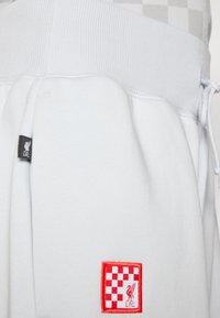 Lyseblå joggebukser med ribbet midje, prydet med en rød firkantet lapp med sjakkmønster og Liverpool Football Club-logo.