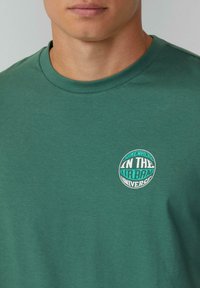 Groene katoenen T-shirt met ronde hals en korte mouwen. Kenmerkt zich door een cirkelvormige grafiek met de tekst "SOMEWHERE IN THE URBAN UNIVERSE" in wit en teal.