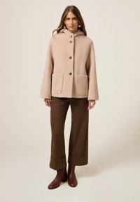 Abrigo beige con capucha y un patrón texturizado, cierre de botones y dos bolsillos frontales, combinado con pantalones de pierna ancha marrones y botas de tobillo burdeos.