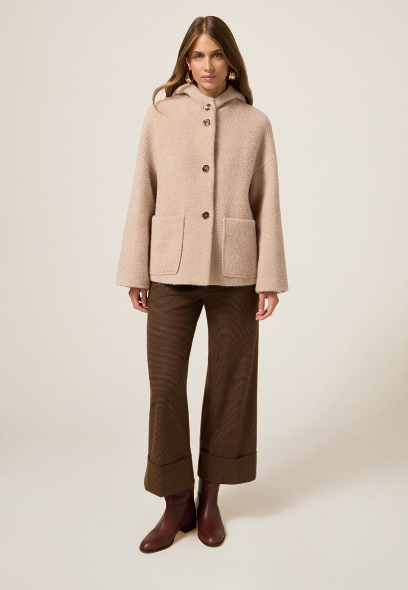 Abrigo beige con capucha y un patrón texturizado, cierre de botones y dos bolsillos frontales, combinado con pantalones de pierna ancha marrones y botas de tobillo burdeos.