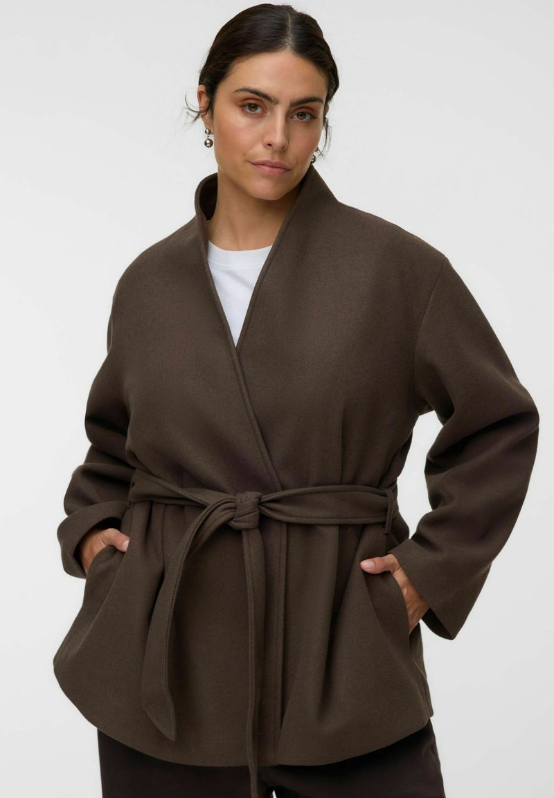 Femme aux cheveux foncés portant un manteau marron ceinturé sur un haut blanc, les mains dans les poches, sur un fond clair uni.