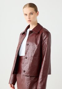 Bordeaux neplederen jasje met een geknipt kraag, knoopsluiting en een voorzak, gecombineerd met een bijpassende rok. Soepele textuur.